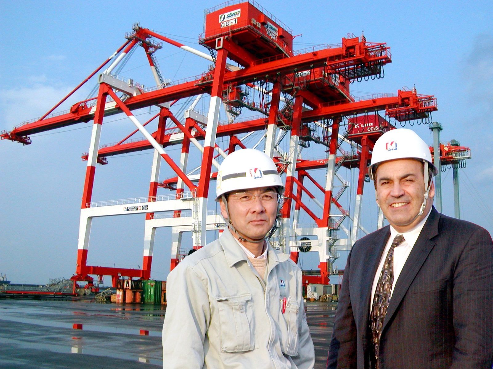 Two men in hard hats standing before red port cranes at a waterfront terminal
