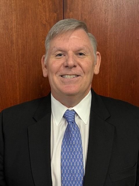 Smiling man in a black suit, white shirt, and blue tie in front of a wood-paneled wall