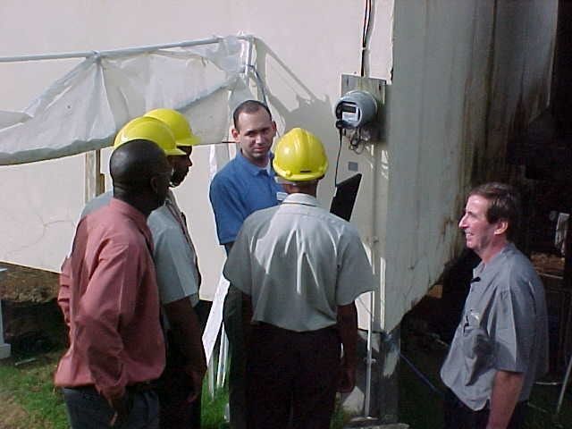 Five workers in hard hats discuss beside industrial equipment outdoors.