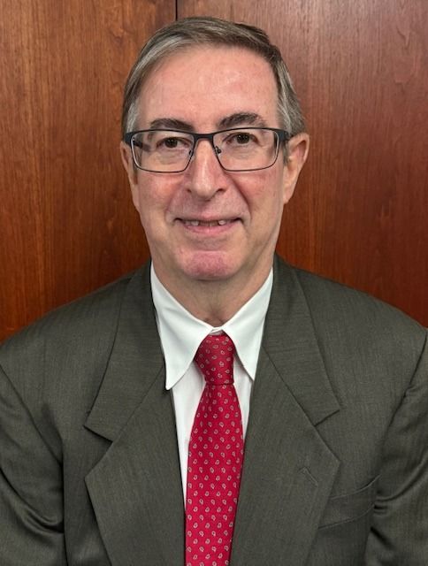 Portrait of a man in a gray suit, white shirt, and red tie against a wood-paneled background