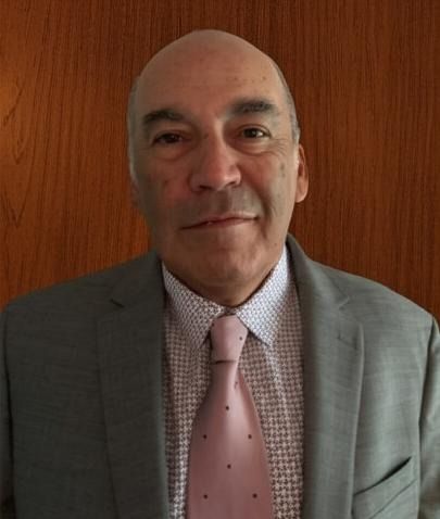 Man in a gray suit and pink tie standing against a wooden wall, looking at the camera.