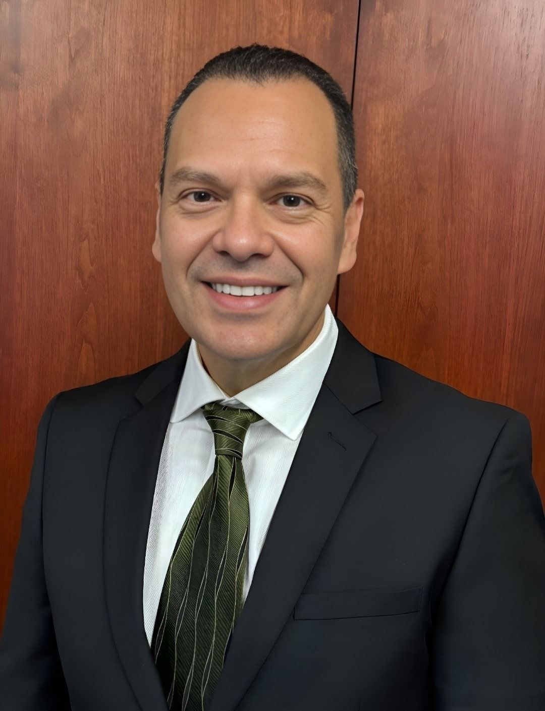Portrait of a smiling man in a dark suit and green tie against a wooden wall.