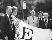 People standing outdoors holding a large white banner with a bold letter “E.”