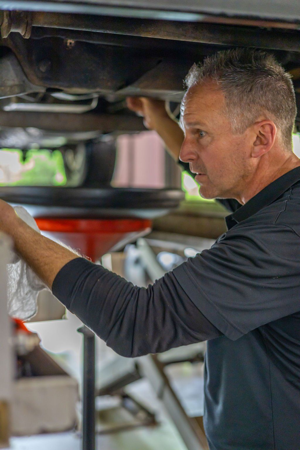 Man changing oil under a car, holding a rag, with a drain funnel in place.