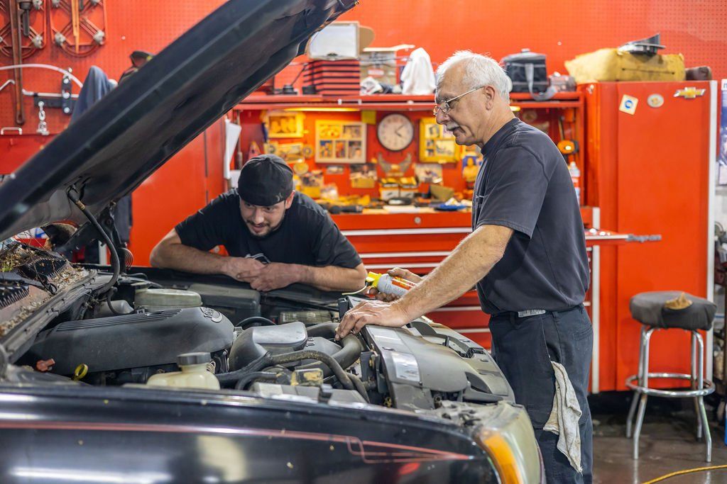 Two mechanics working on a car engine in a garage. One looks inside, the other leans in and observes.