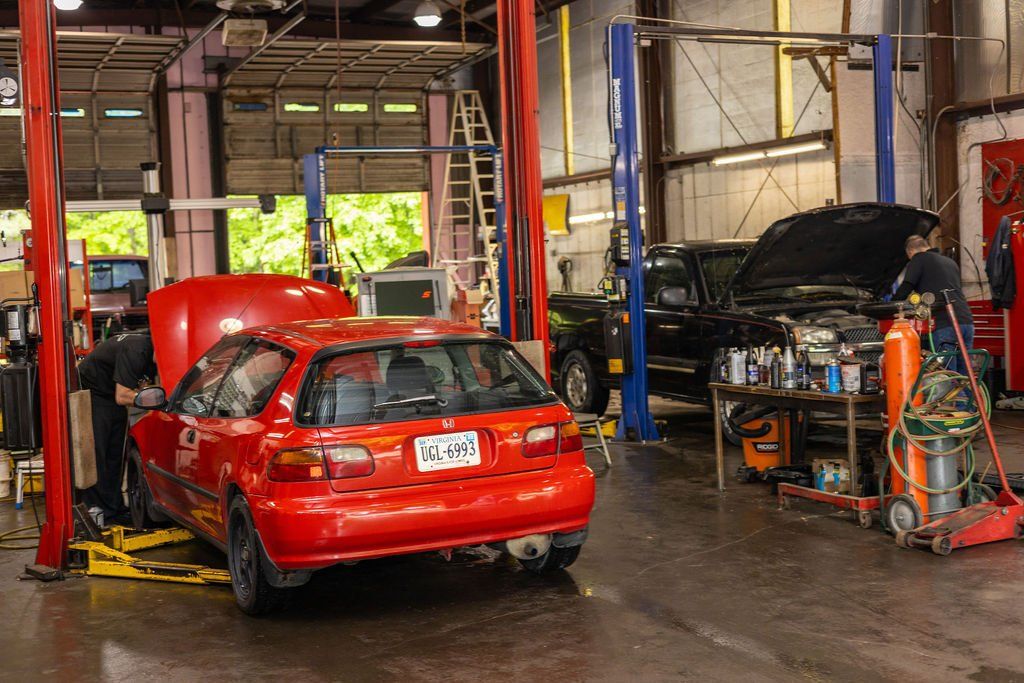 Red car on lift being worked on inside a garage. Another vehicle with an open hood is in the background.