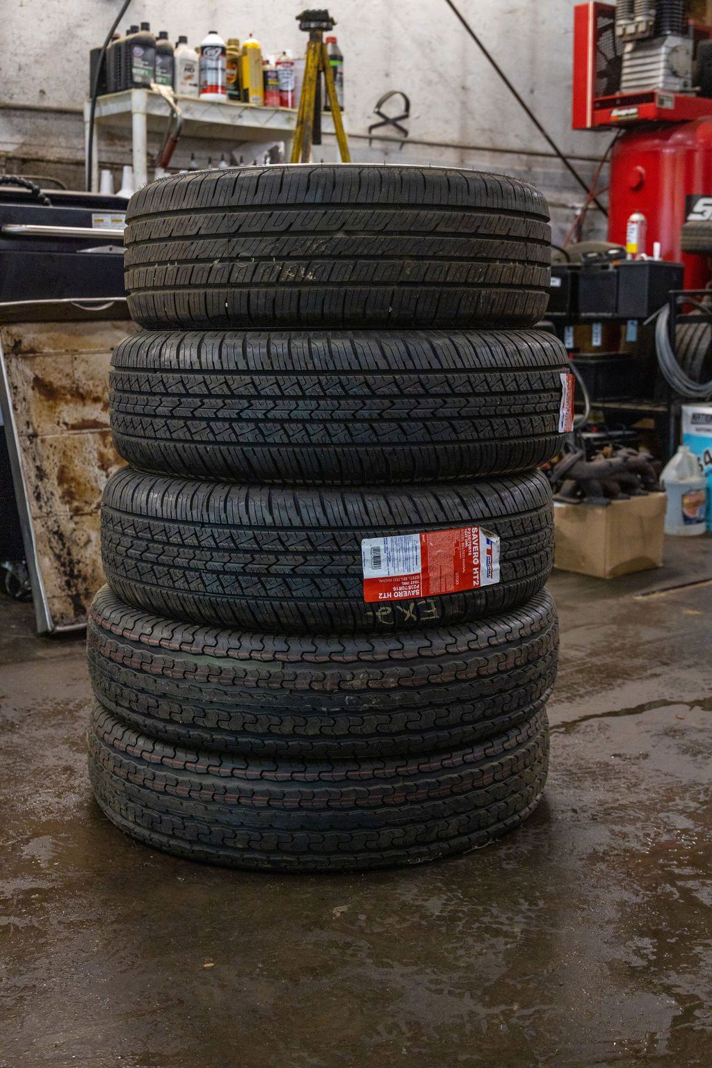 Four stacked tires with black treads in a garage setting, one tire has a red and white label.