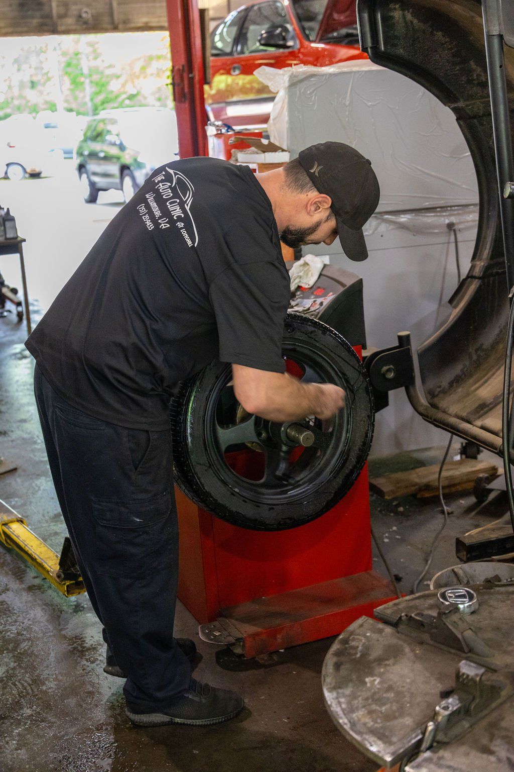 Mechanic balancing a tire in a garage. He wears a black shirt and cap while working on the wheel.