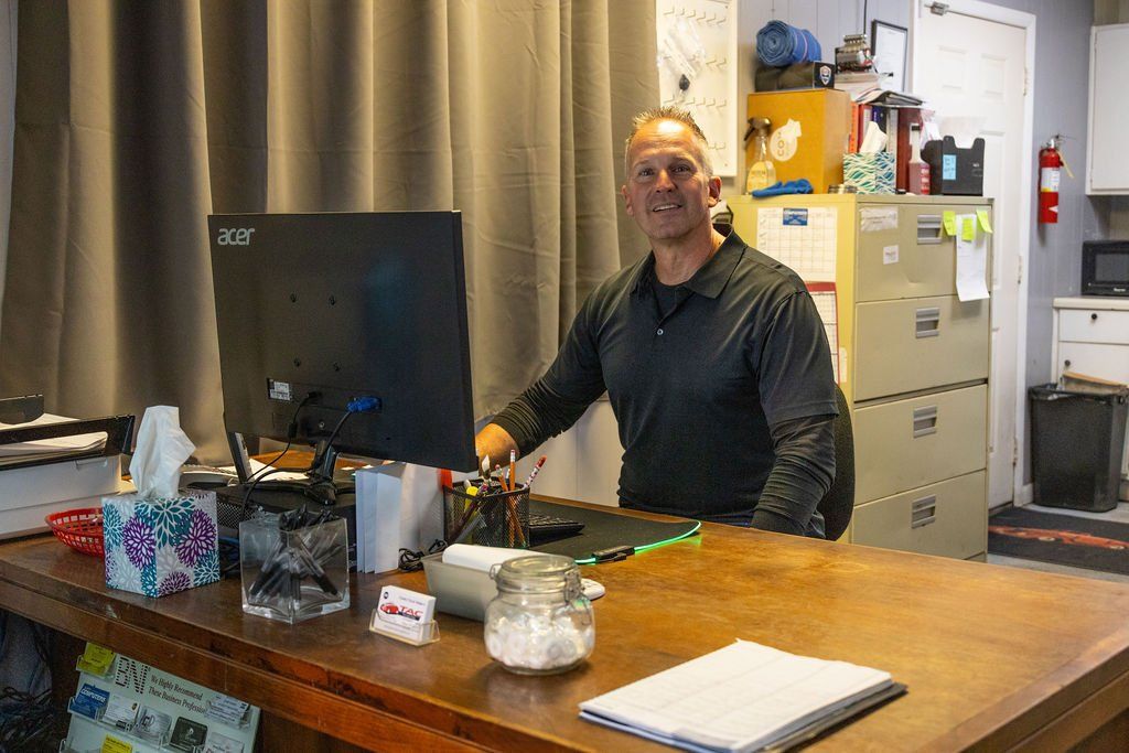 Man behind desk, smiling, in office setting with computer and filing cabinet.
