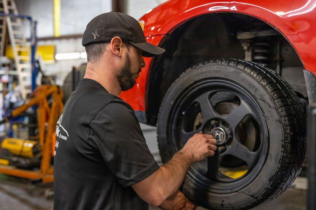 Mechanic installing a car tire in a garage. The car is red, and the mechanic wears a black cap and shirt.