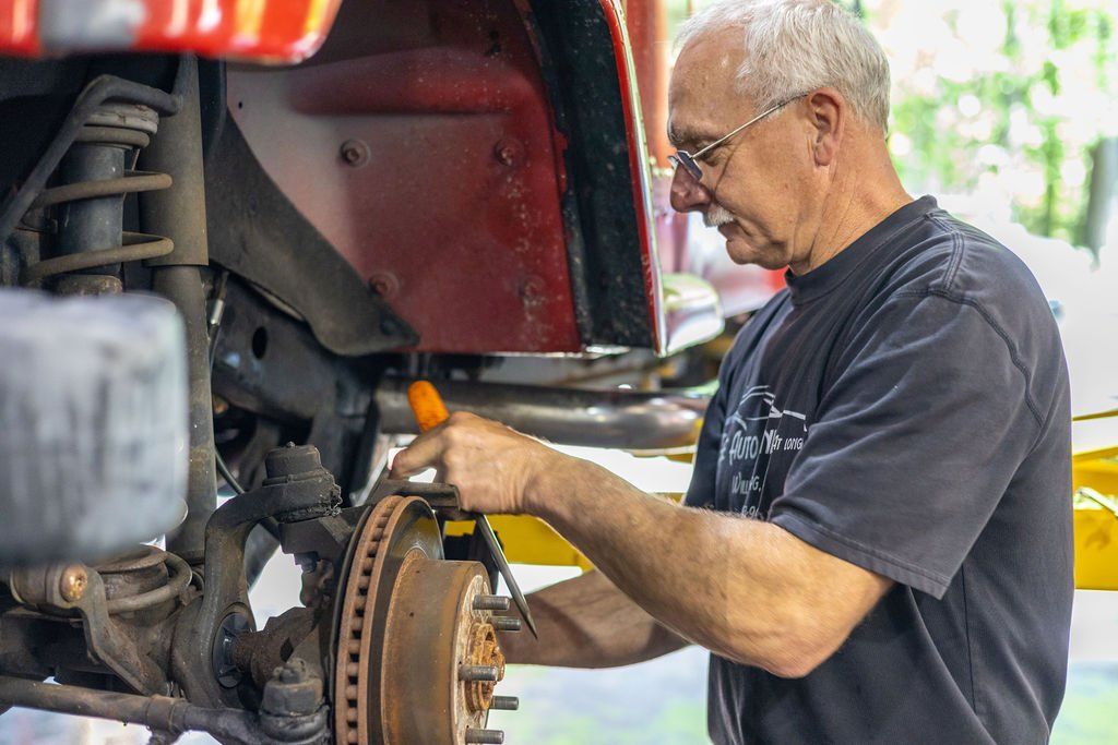 Mechanic working on car brakes; red vehicle. Man with glasses holds tool, examining parts in a garage setting.