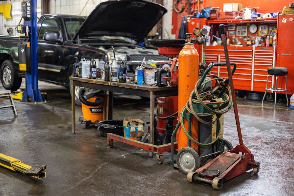 Inside auto repair shop: black pickup truck with open hood, workbench with tools, welding equipment, and floor jack.