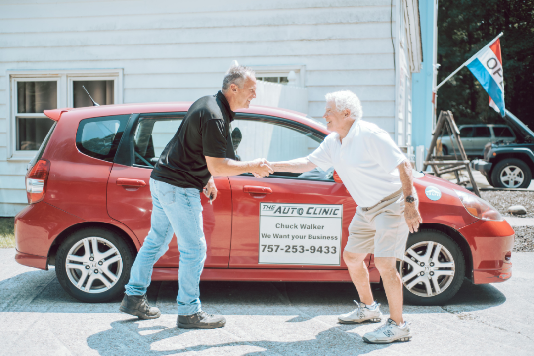 Two men shaking hands in front of a red car, near a building with an auto clinic sign.