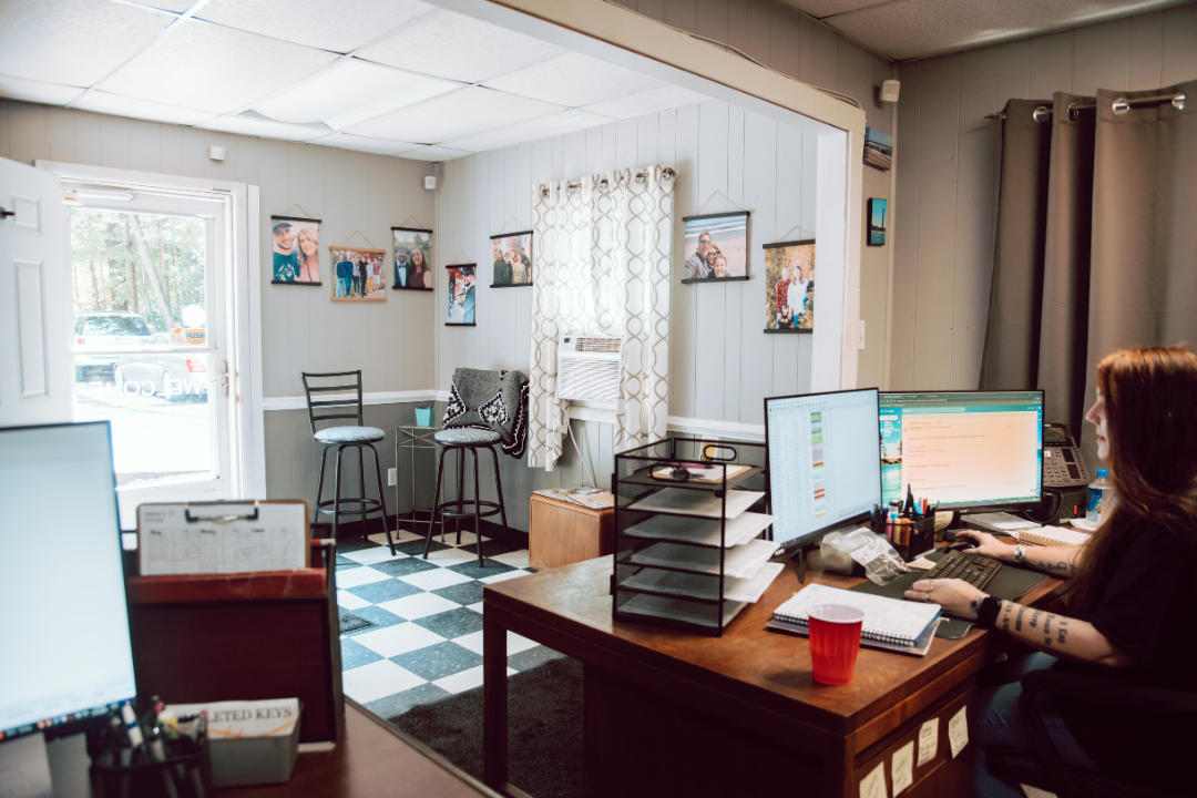 Office interior with a person at a desk using a computer. Black and white checkered floor, photos on the wall.