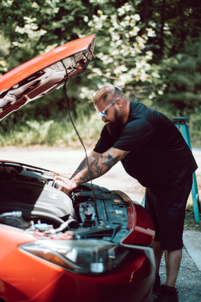 Man with tattoos examining engine of a red car outdoors, hood open.