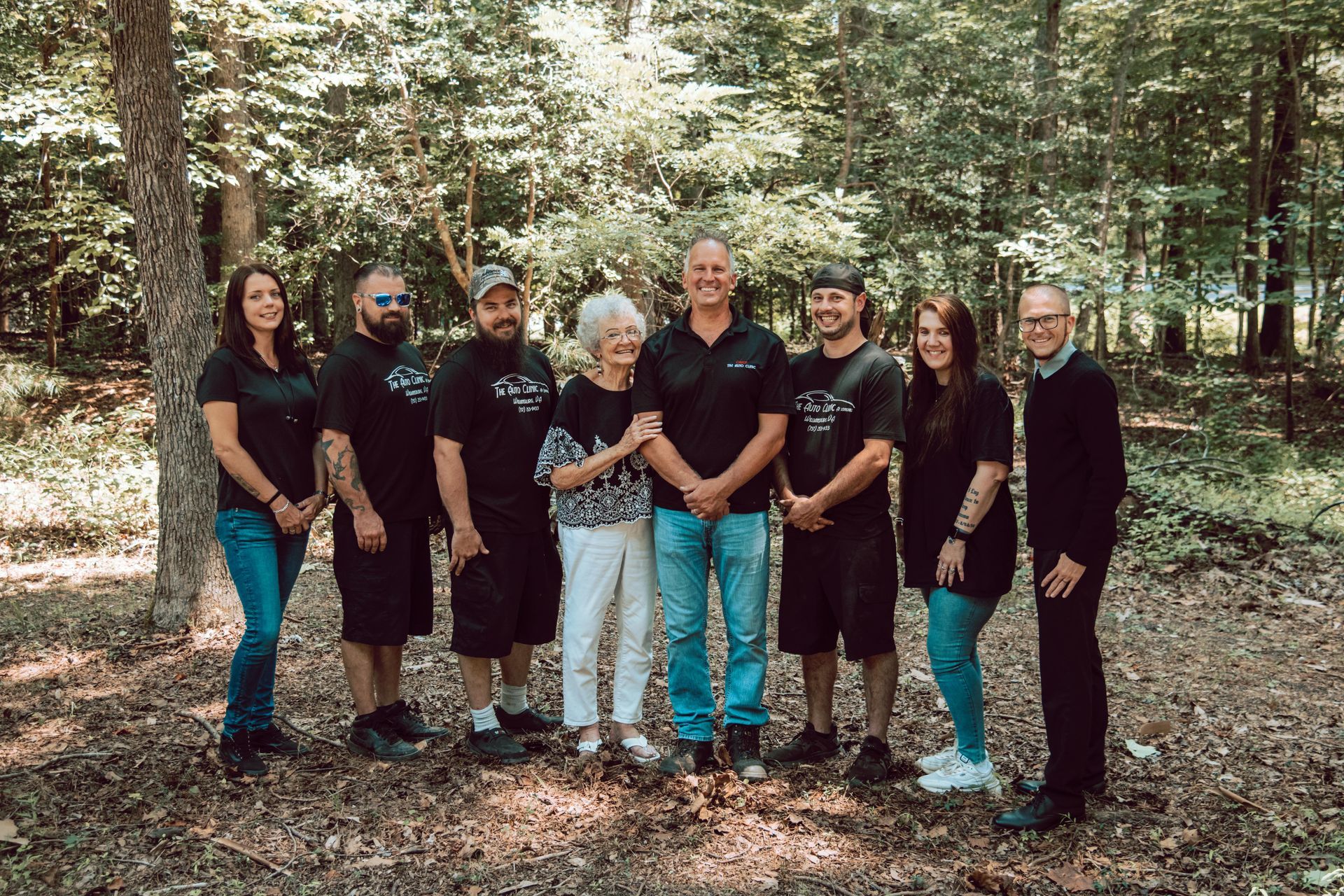 Group of eight people standing outdoors in front of trees; wearing casual clothes.