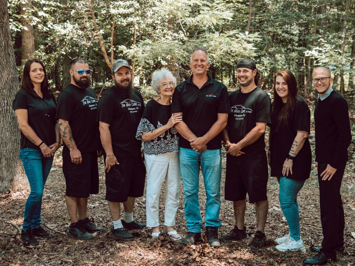 Group of eight people posing in front of trees. Some wearing black shirts and jeans. Older woman has arm around a man.