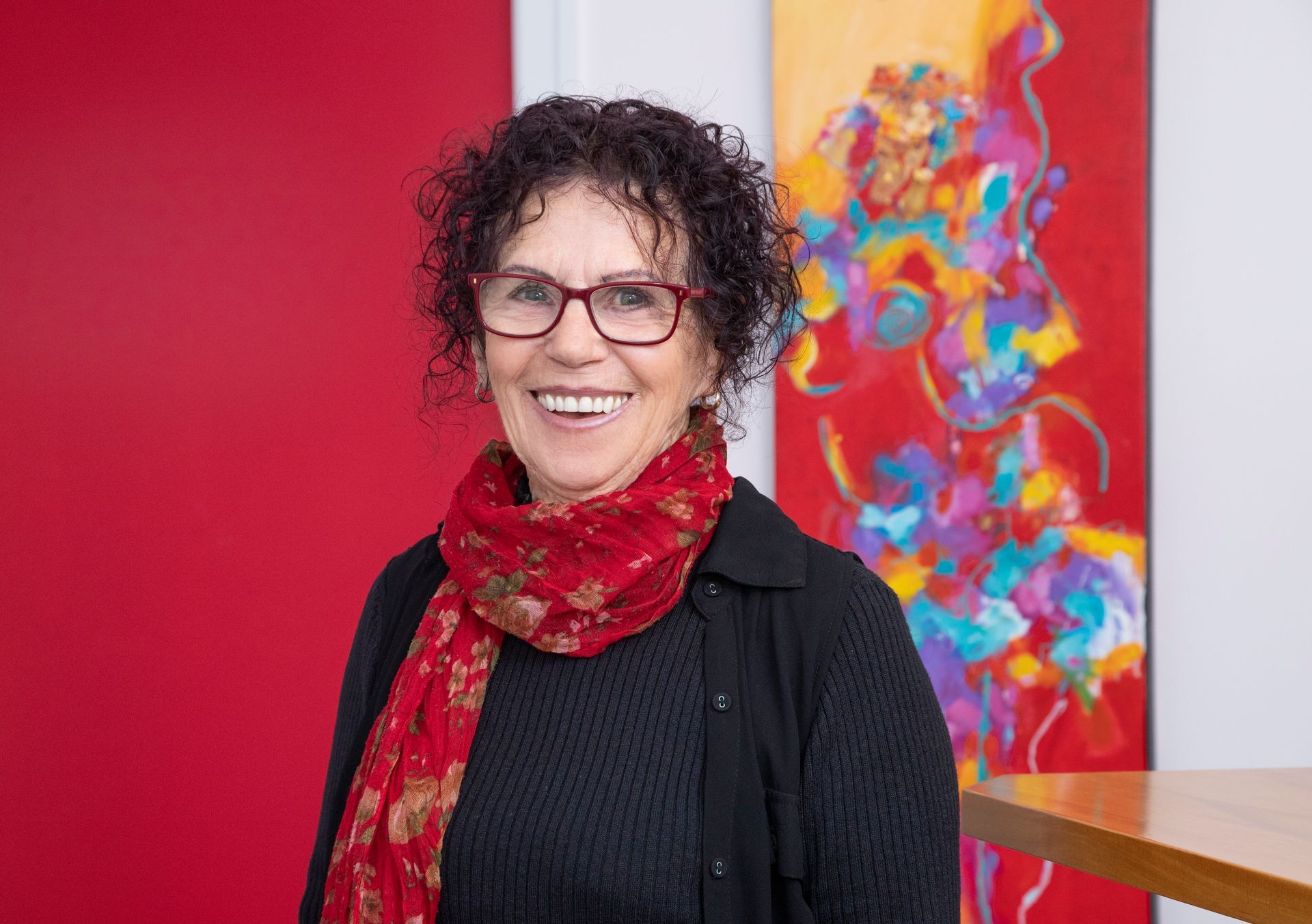A woman wearing glasses and a red scarf is smiling in front of a painting.