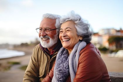 Smiling couple sitting outdoors by the water, wearing warm jackets on a sunny day
