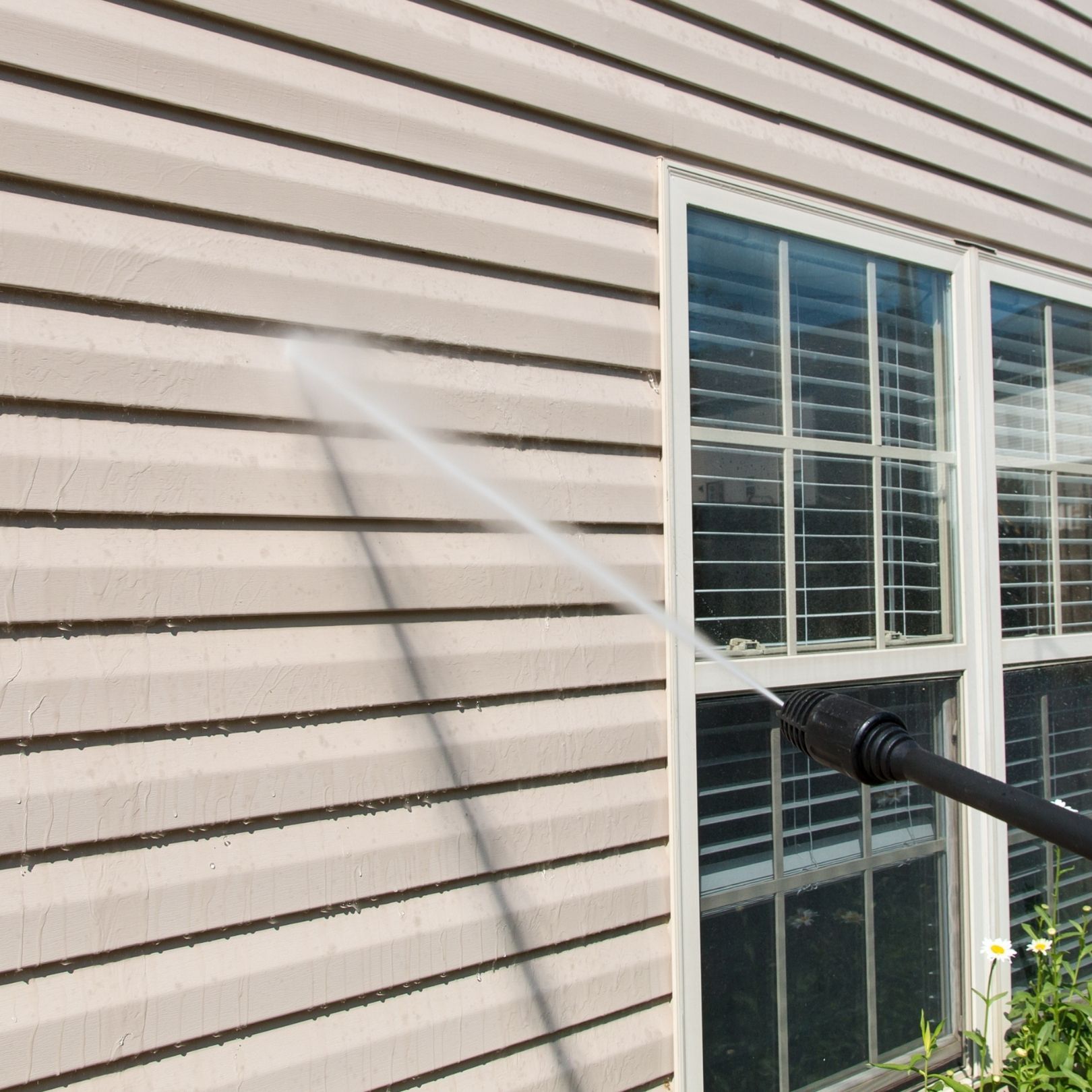 Garden hose spraying water on beige house siding beside a window