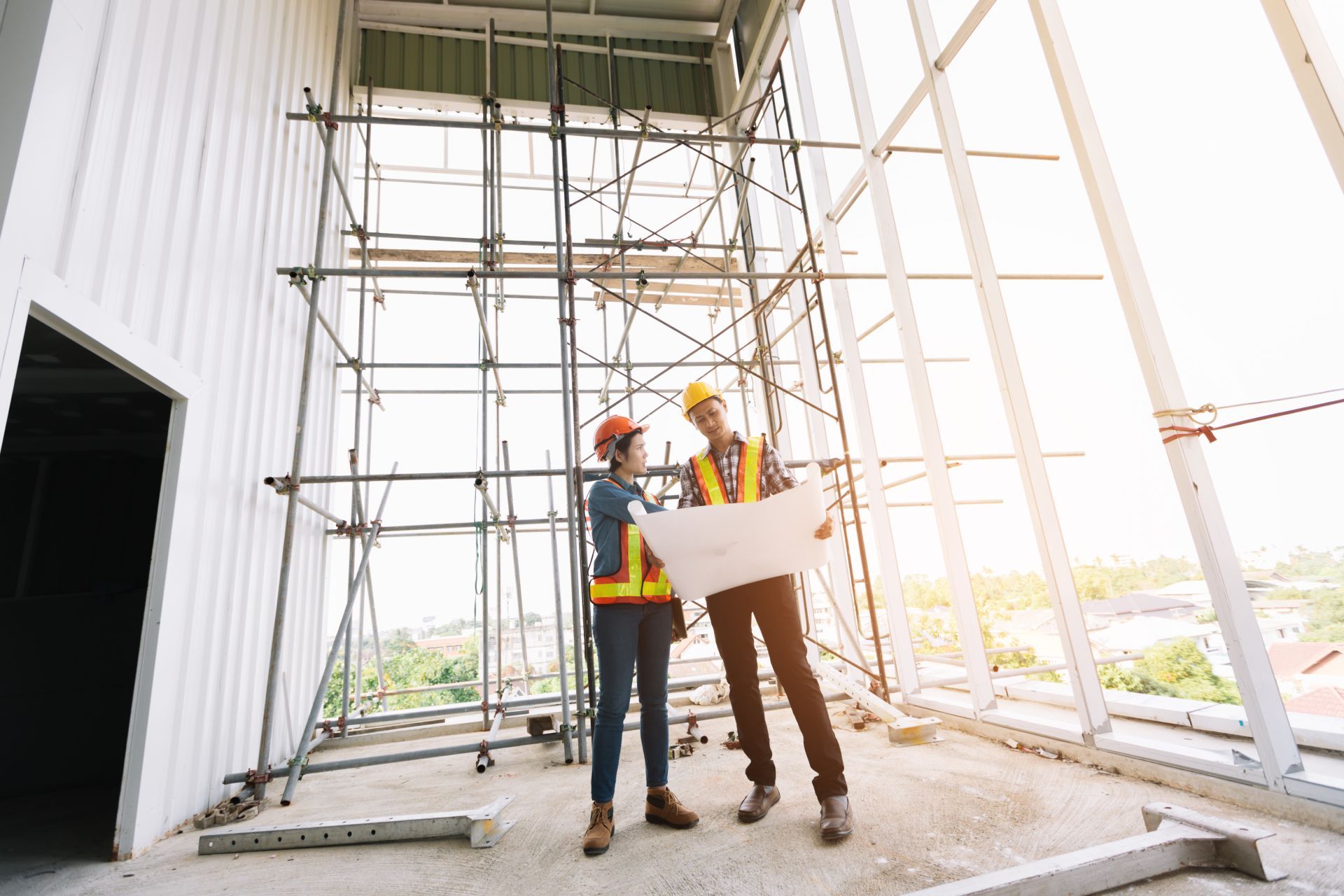 Two construction workers in hard hats reviewing plans at a scaffolding site inside a steel-framed building