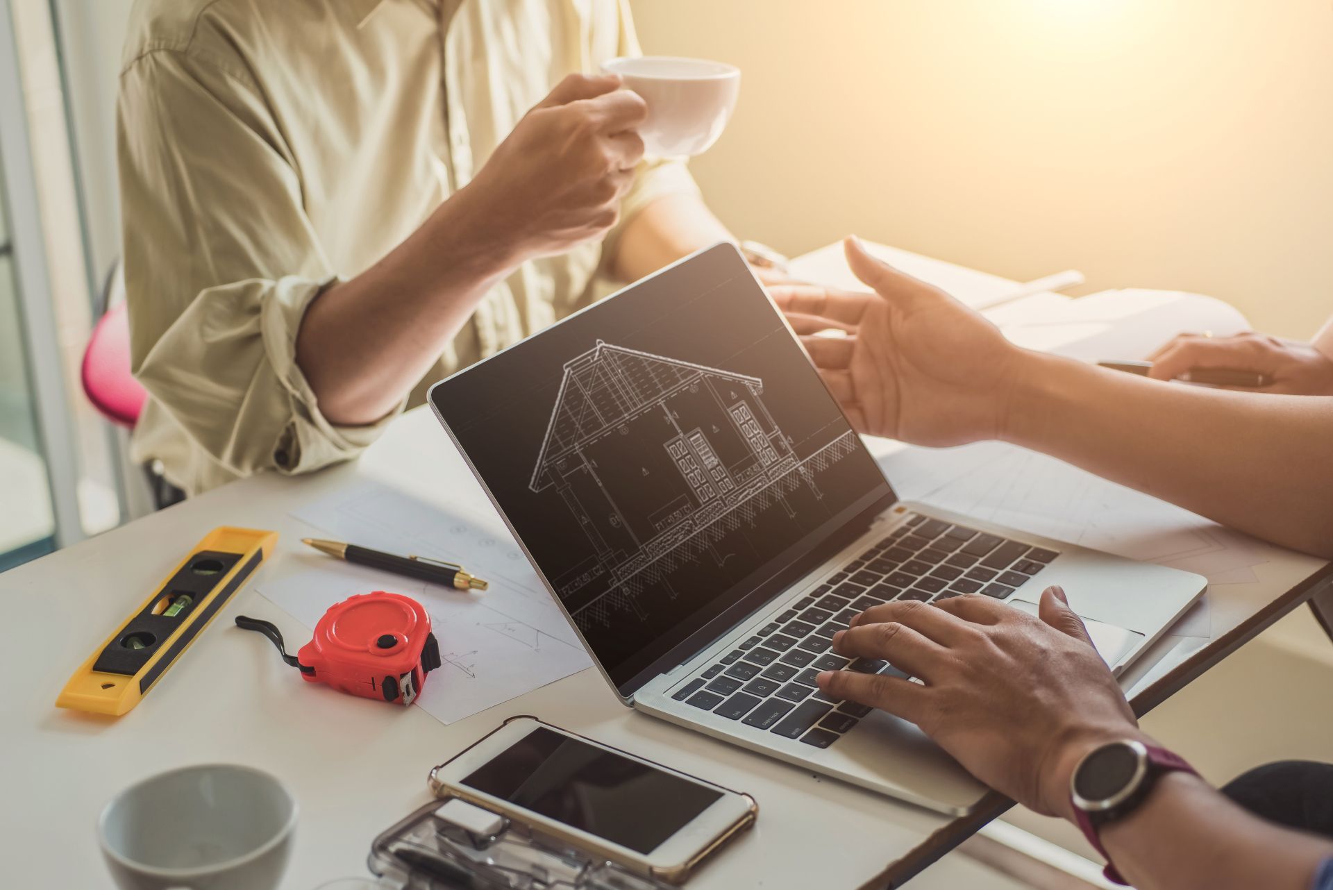 Hands working on a laptop with a house plan on screen, surrounded by measuring tools and a coffee mug.