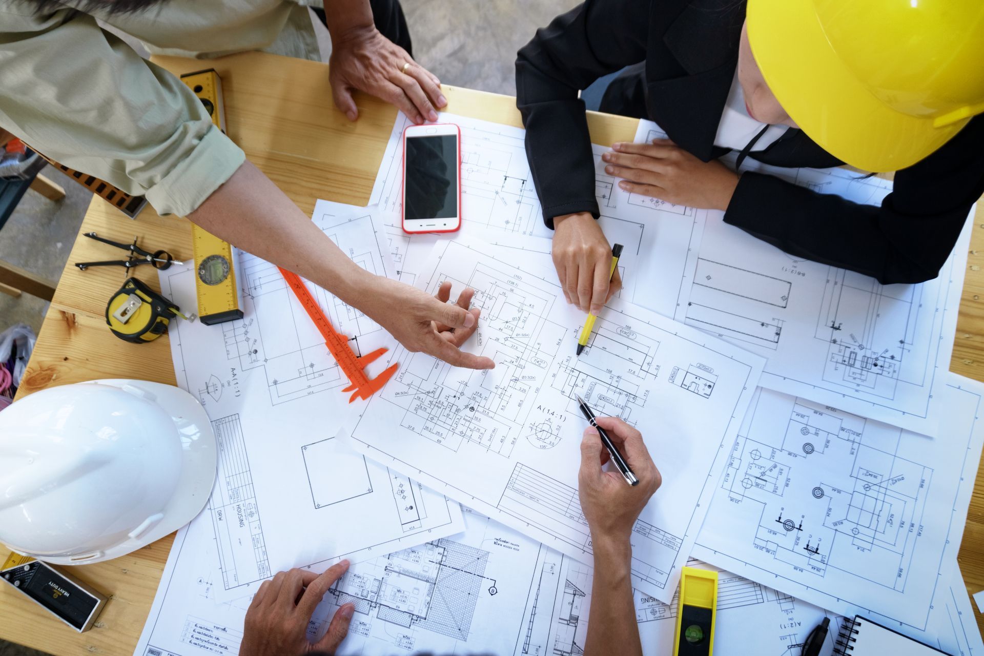 Engineers reviewing blueprints around a table, with hard hats, tools, and a smartphone.