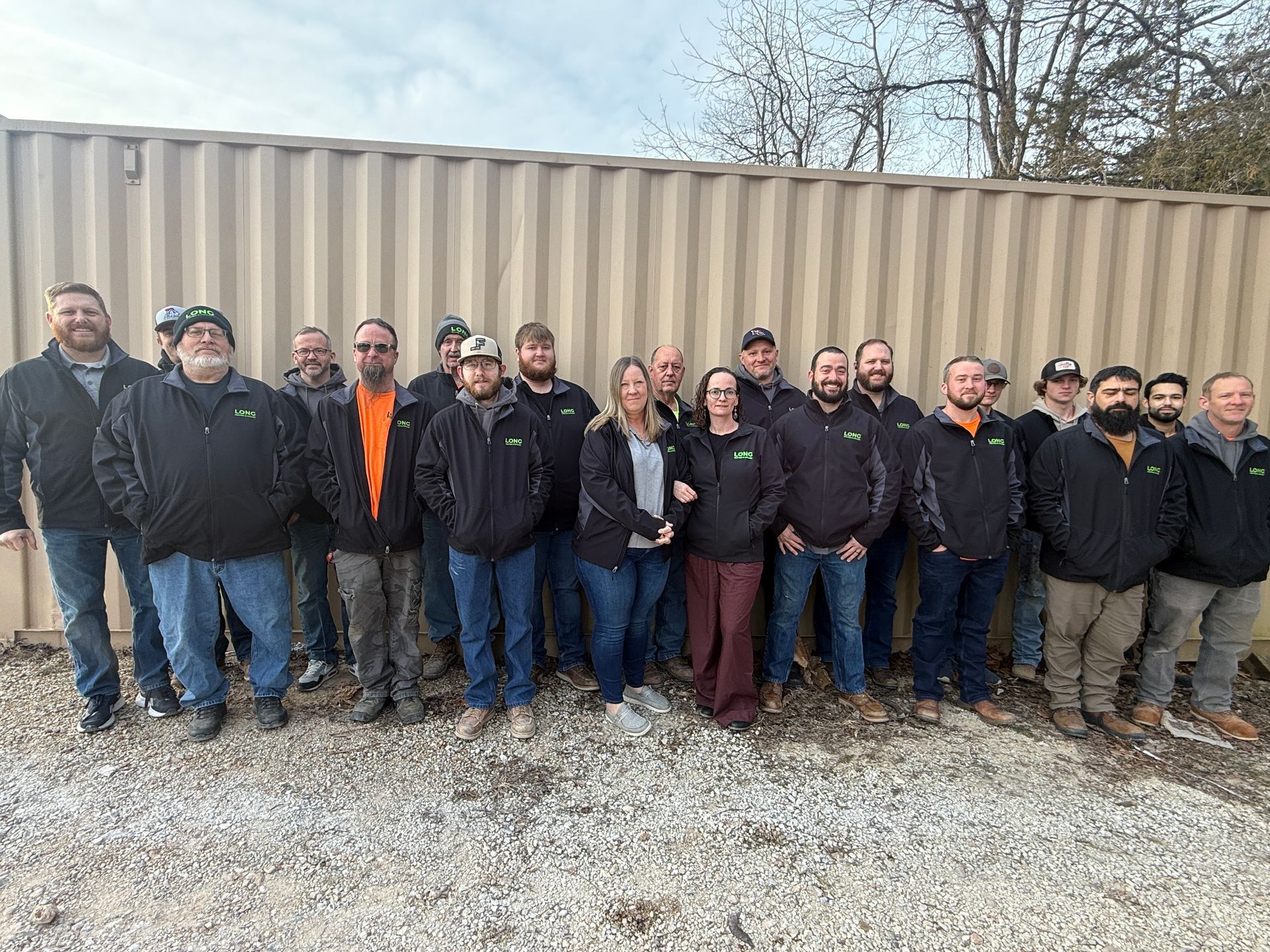 Group of workers standing in front of a tan shipping container on a gravel lot, wearing dark jackets.