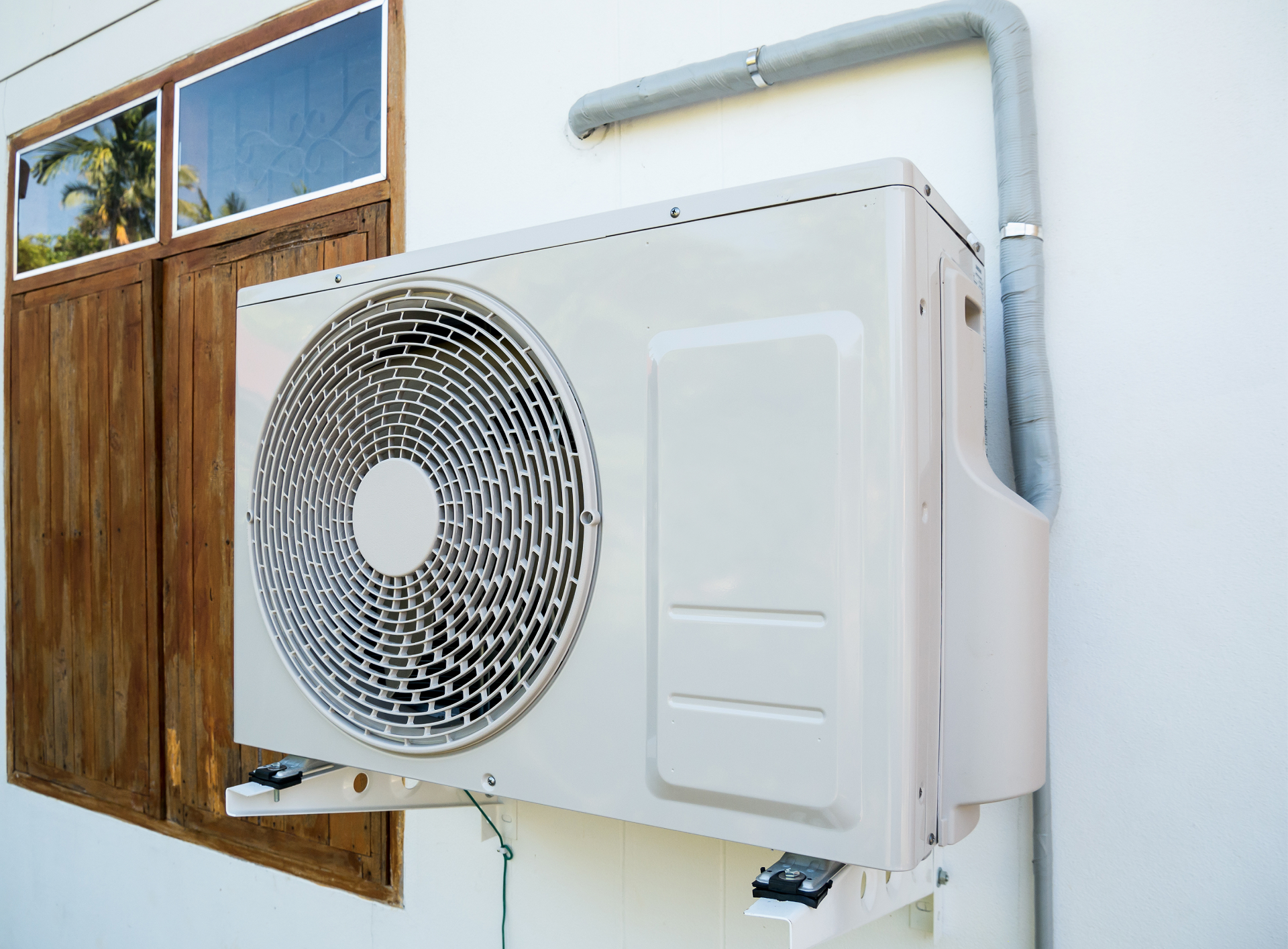 Outdoor wall-mounted air conditioner unit beside a wooden window shutter and white pipe on a building wall