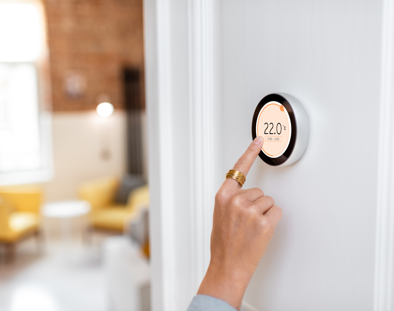 Hand pressing a round digital thermostat on a white wall, with a blurred living room in the background