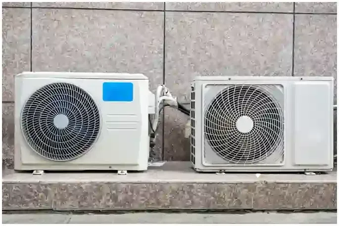 Two white outdoor air-conditioning units on a tiled floor against a gray wall, connected by tubing