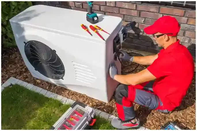Technician in red uniform repairs an outdoor HVAC unit beside a brick wall, with tools on top.