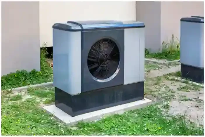 Outdoor air-conditioning unit beside a building, gray and black on a concrete pad, surrounded by grass.
