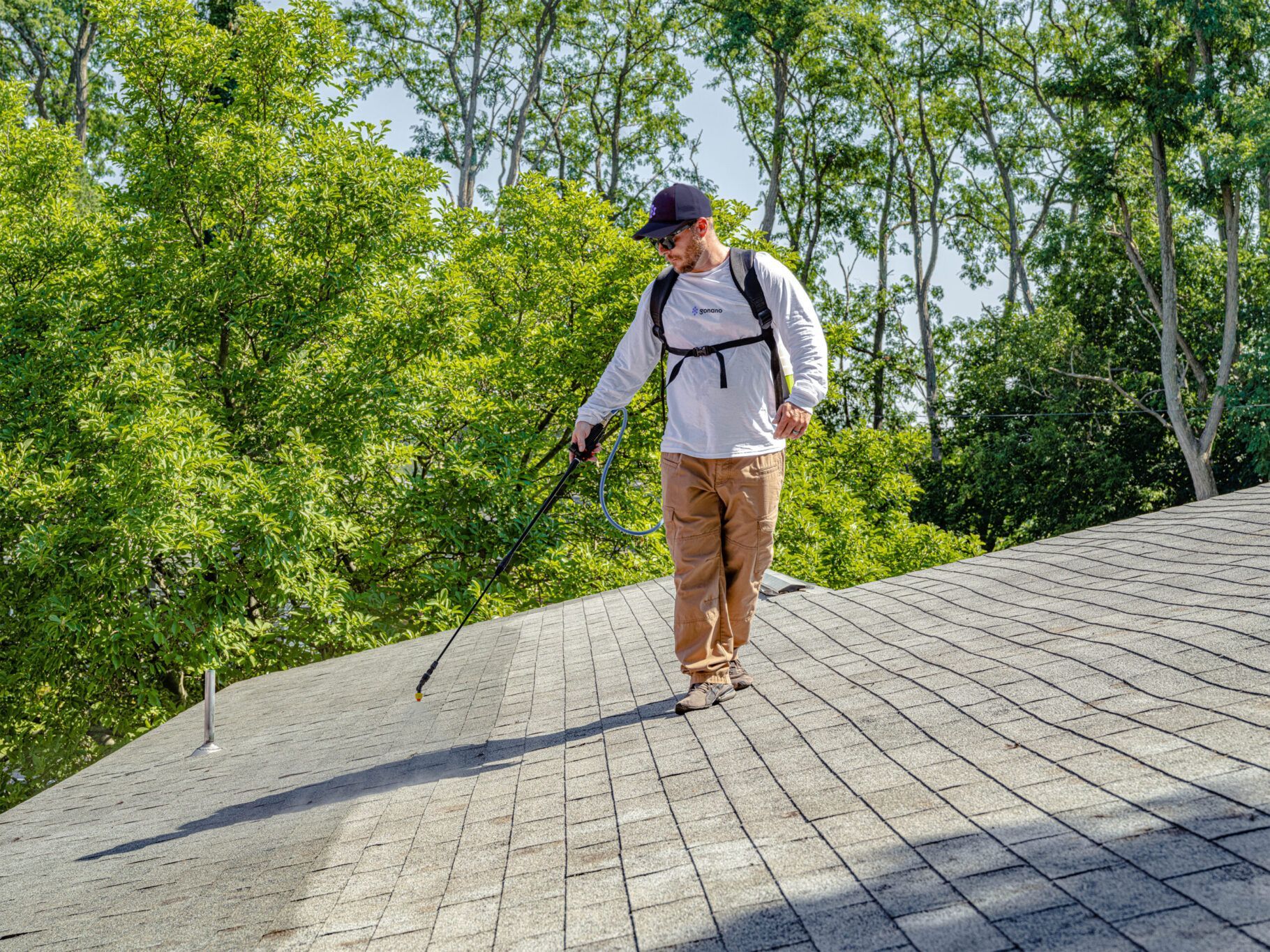 Person spraying a roof with a backpack sprayer; green trees in the background.