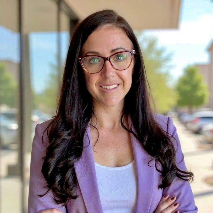 Woman in glasses and a lavender blazer smiling outdoors near a building