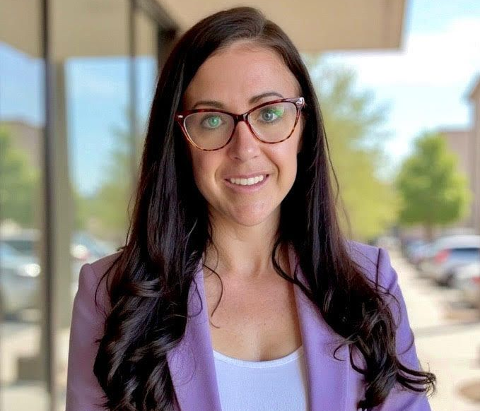 Smiling person with glasses and long dark hair in a lavender blazer standing outdoors near a building