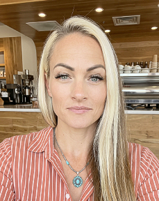 Portrait of a blonde woman in a striped shirt, seated in a wood-paneled café with coffee equipment behind her