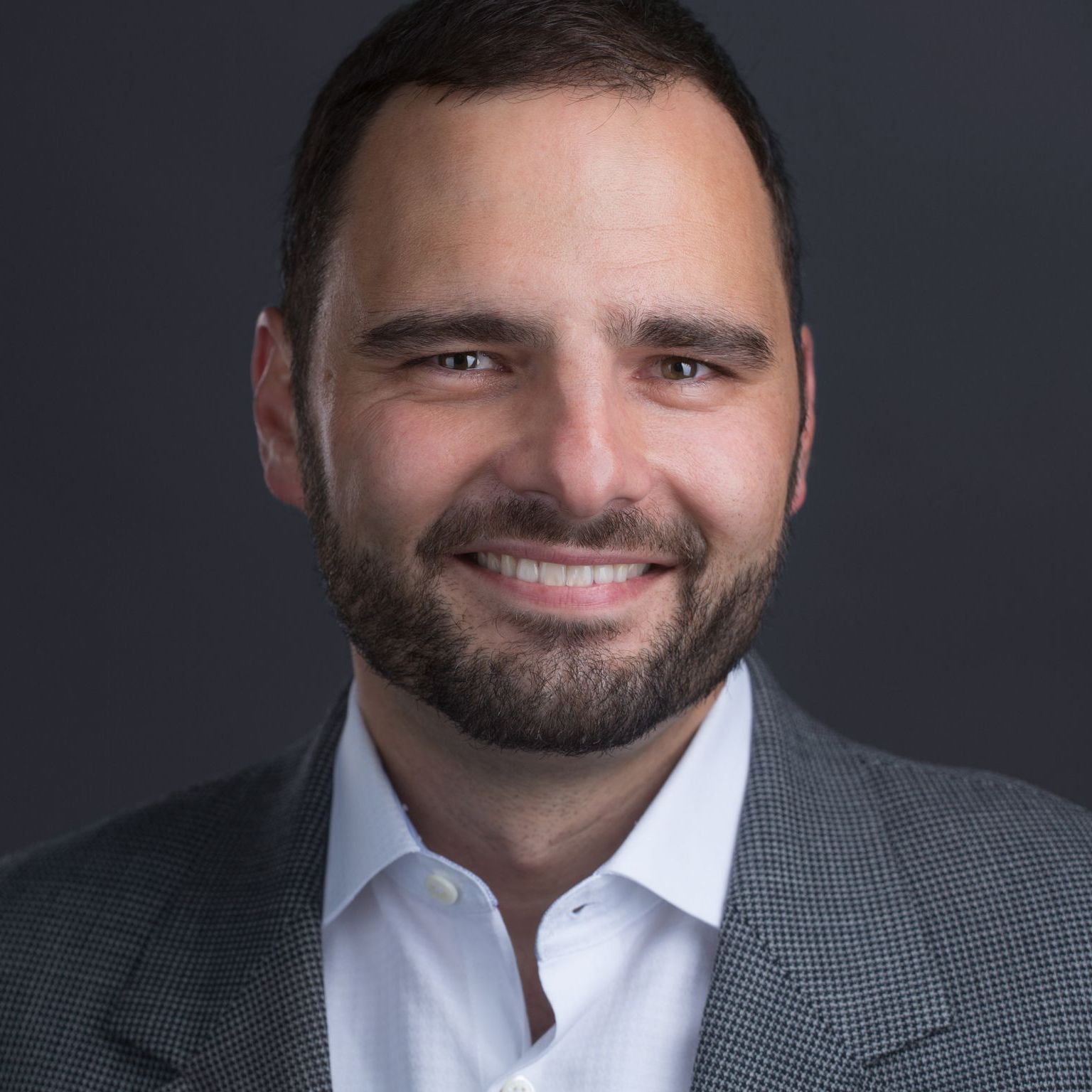 Studio headshot of a smiling man in a dark blazer and white shirt against a gray background