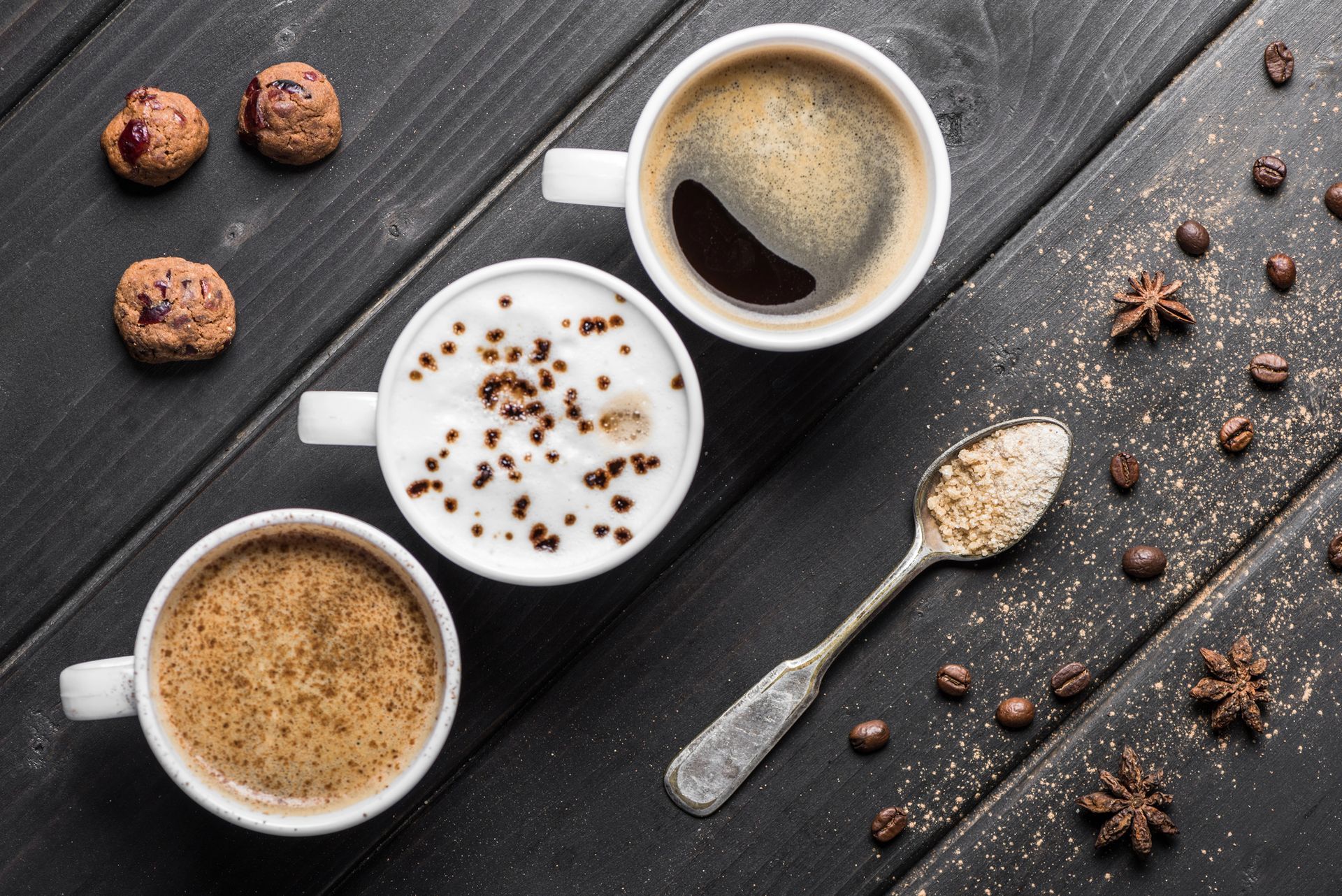 Tre tazze da caffè, biscotti, un cucchiaio con lo zucchero e chicchi di caffè su un tavolo di legno nero.