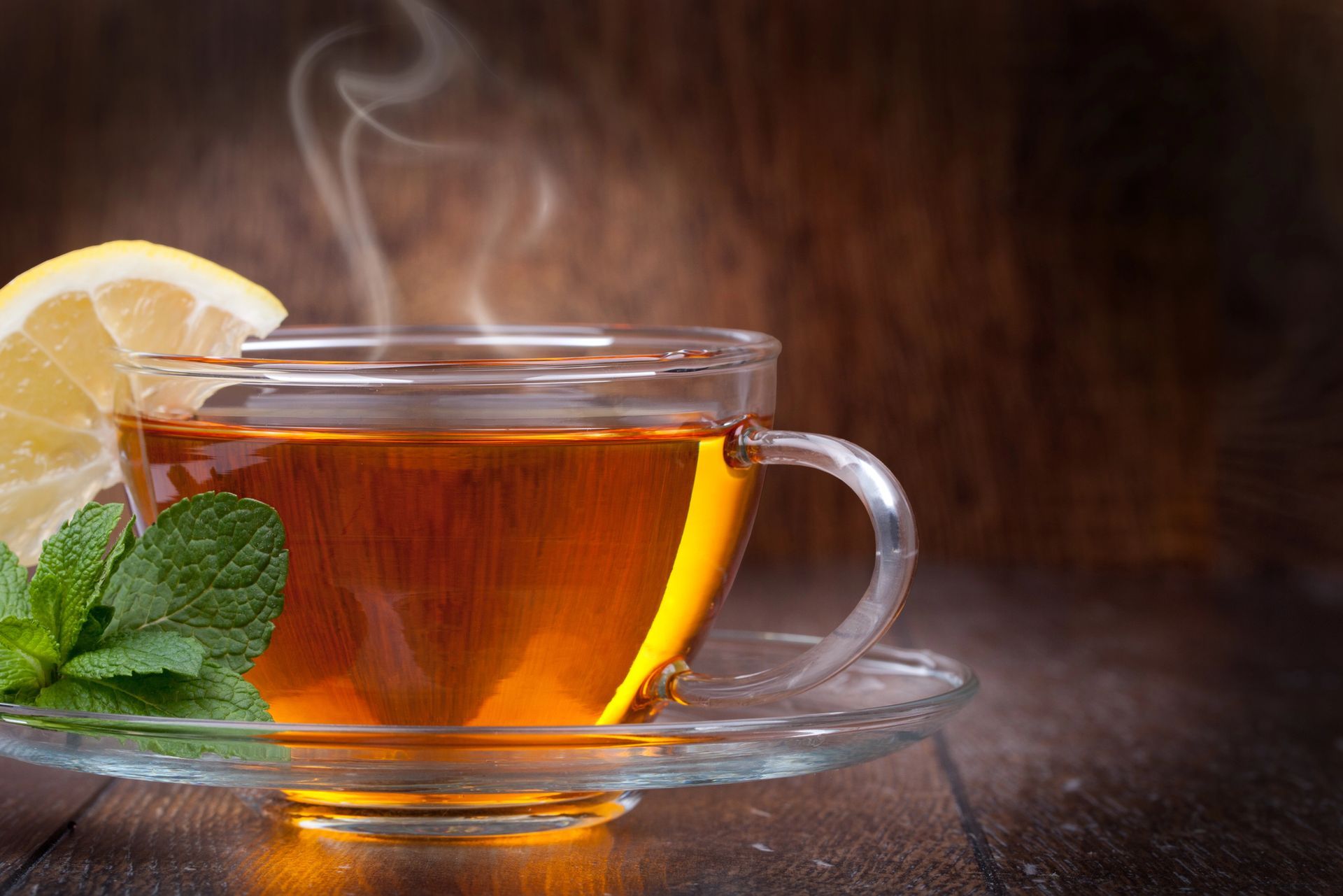 Tazza di tè fumante con fetta di limone e foglia di menta su un piattino, sfondo di legno scuro.