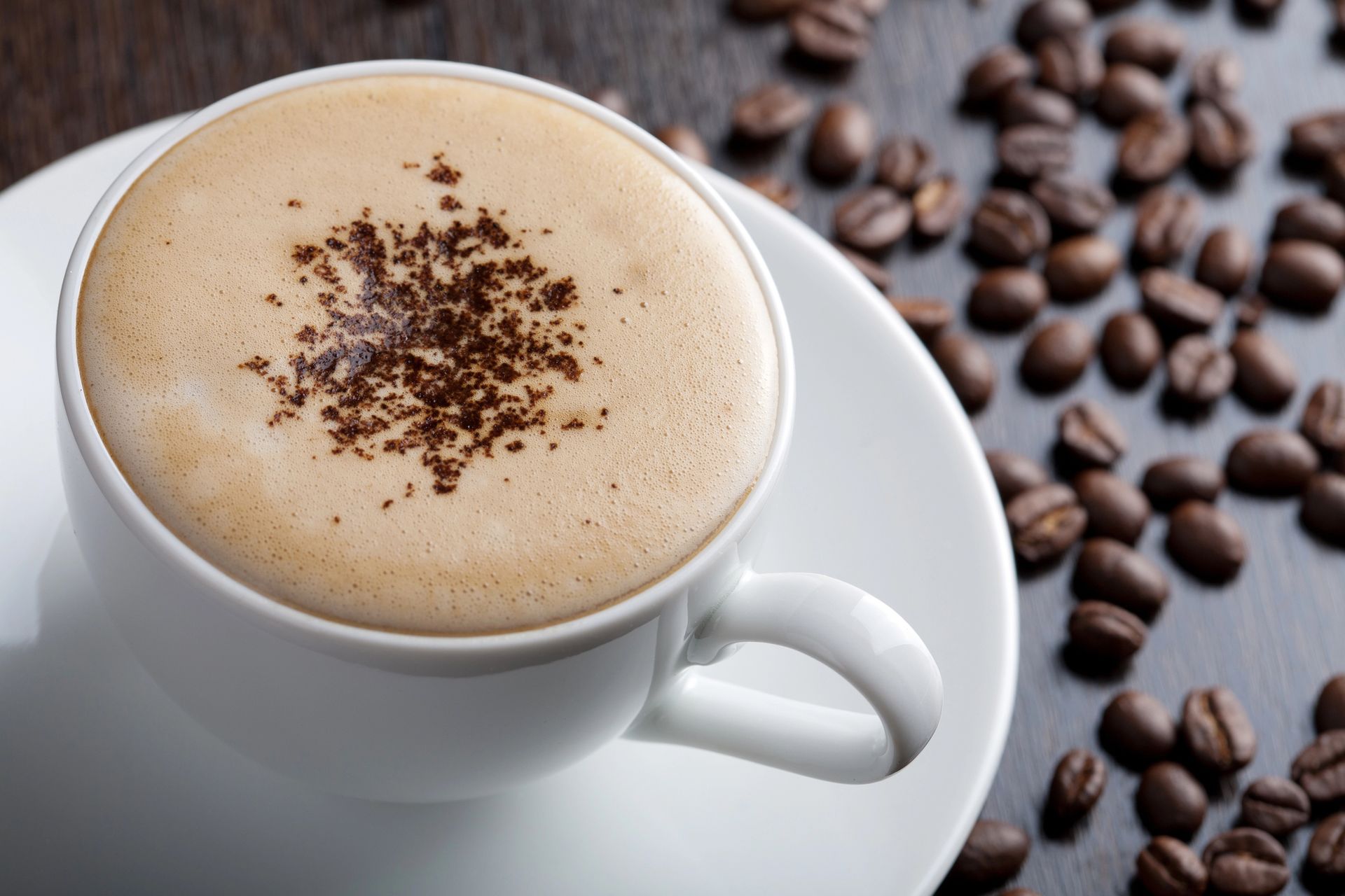 Cappuccino in tazza bianca su piattino, cosparso di cacao, accanto a chicchi di caffè sparsi.