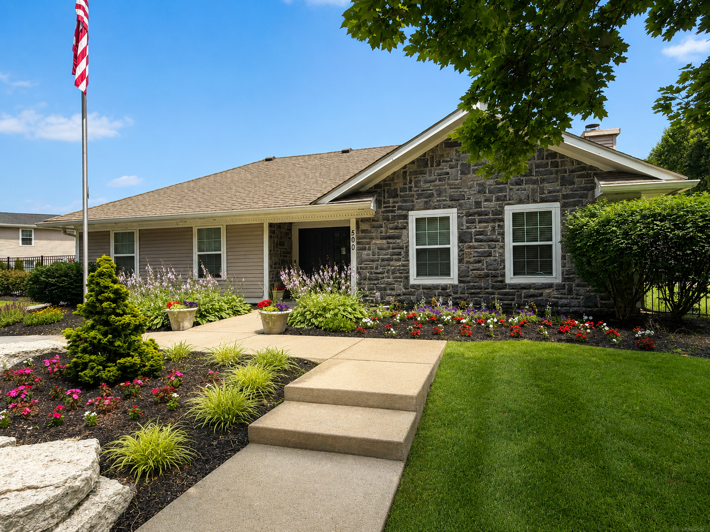 Single-story house with stone facade, manicured lawn, flower beds, and a flagpole in the front yard
