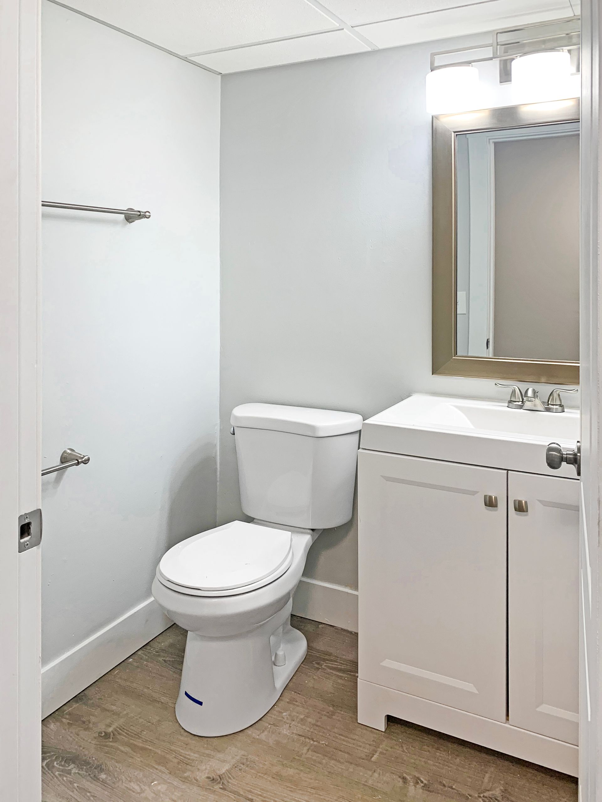 Small white bathroom with toilet, vanity, mirror, and shower curtain rod, viewed from the doorway