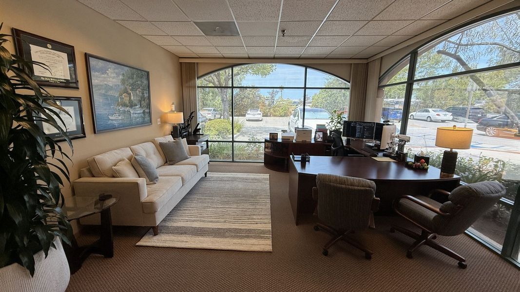 Bright office lounge with sofa, desk, chairs, and large windows overlooking a parking lot