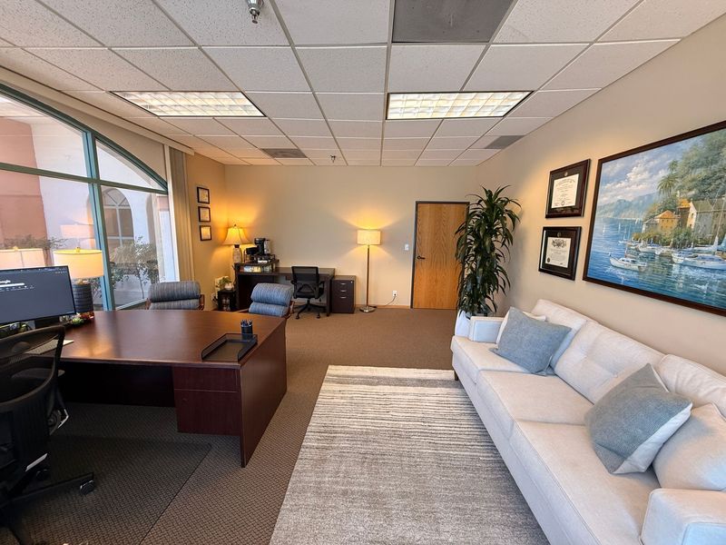 Modern office lounge with desk, sofa, artwork, and large window in a bright carpeted room