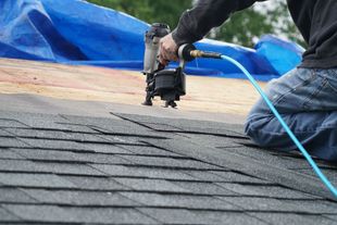 A roofer uses a pneumatic nail gun to install black shingles on a plywood roof deck with a blue tarp in the background.