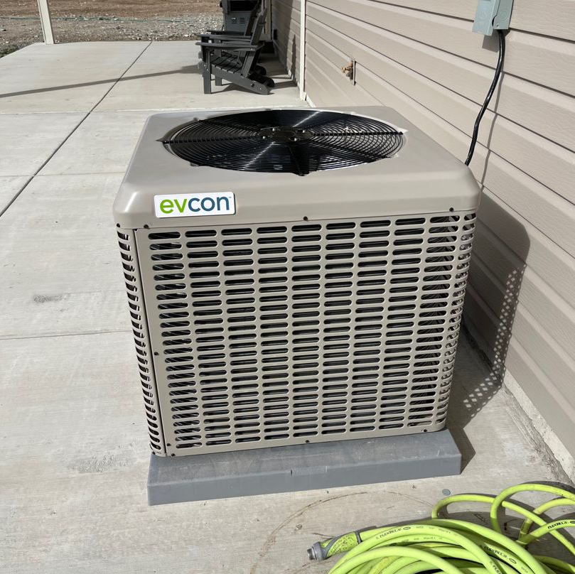 Two outdoor air conditioning units beside a house, next to a stone retaining wall and grass.