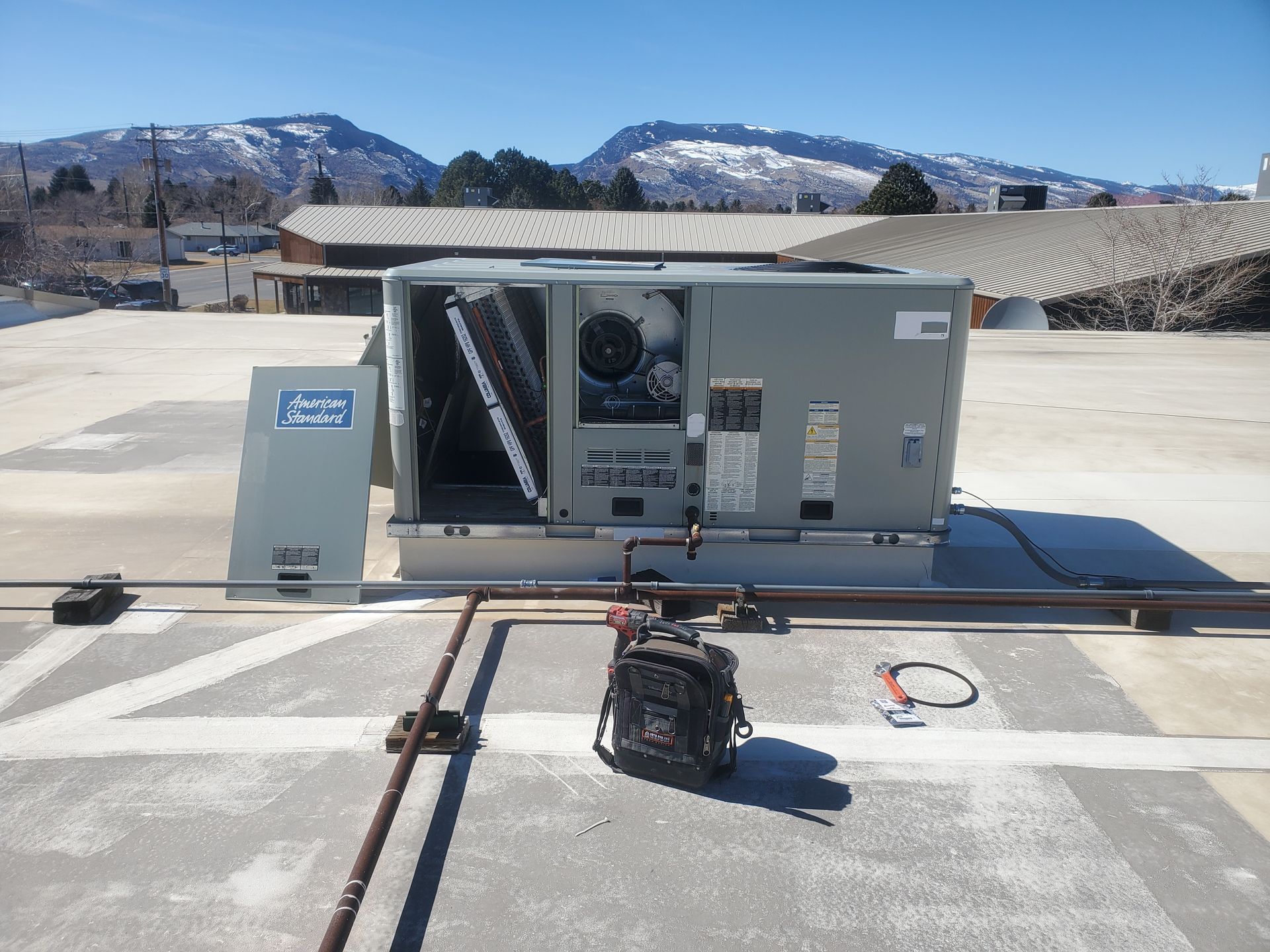 Outdoor HVAC unit with open panel, exposed wiring, and a tool bag on a rooftop under a clear blue sky.