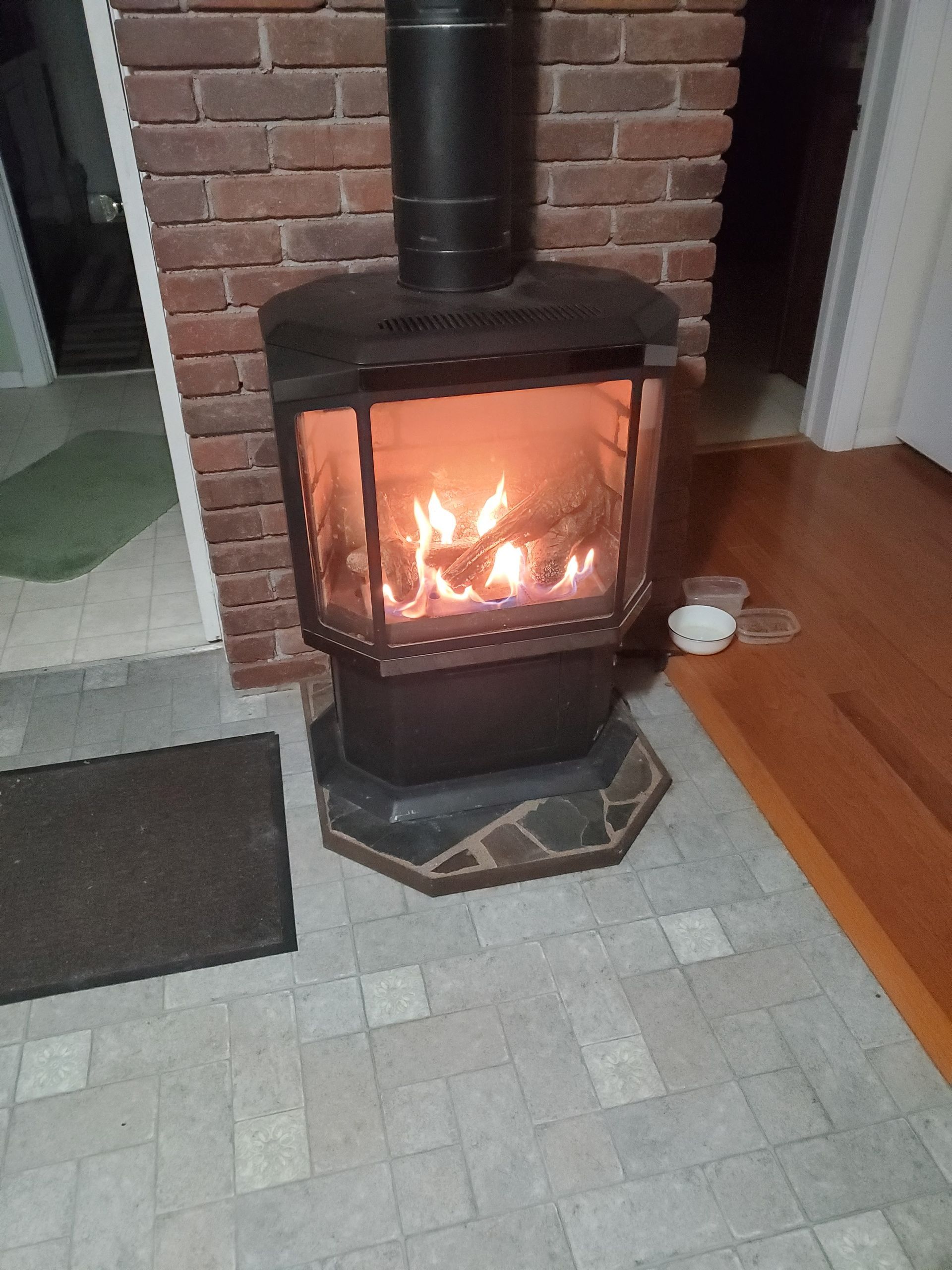 Lit wood-burning stove with black chimney in a brick fireplace, in a living room corner.