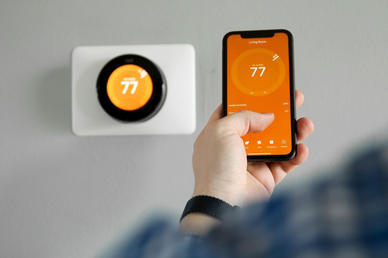Hand holding a phone controlling a wall thermostat, both showing 77° on orange screens
